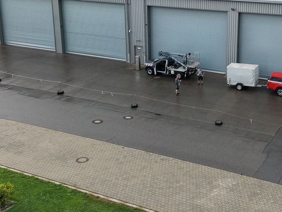Nasse Asphalt- und Pflasterfläche vor einer Halle, darauf ein Hubsteigerfahrzeug mit Anhänger, von oben aufgenommen