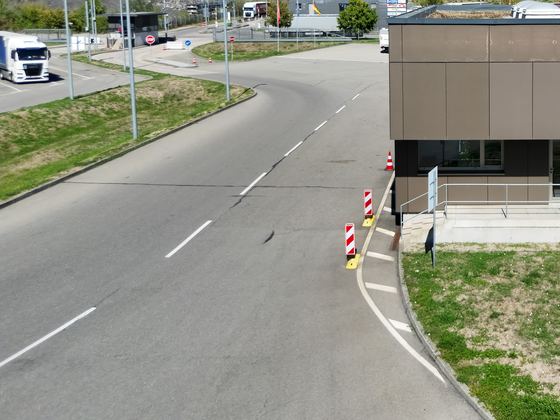 Geschwungene Zufahrtsstraße mit Bordstein und Leitbaken vor einem Pförtnerhaus, von oben aufgenommen